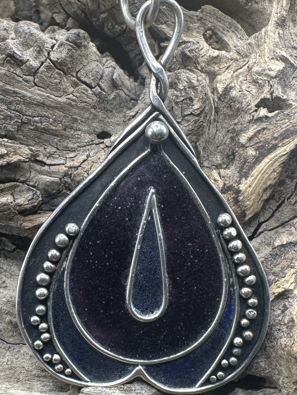 Unique vulva sculpted silver pendant with dark violet and dark blue violet fused glass displayed on driftwood.